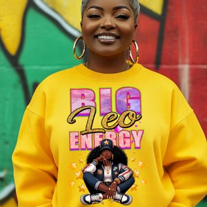 May include: Yellow sweatshirt with the text "BIG Leo ENERGY" in pink and gold lettering. A cartoon illustration of a woman with a star on her hat, wearing a blue jacket, jeans, and sneakers, is sitting with her legs crossed.