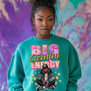 Op de afbeelding: Een turquoiseblauw sweatshirt met de tekst "BIG Gemini ENERGY" in roze en gouden glitterletters. Een cartoonillustratie van een vrouw met een roze muts en een gouden ketting zit in het midden van het sweatshirt.