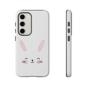 May include: A white phone case featuring a cute bunny face design. The bunny has pink ears, blush, and black whiskers and eyes. The case has a black border and a camera cutout.