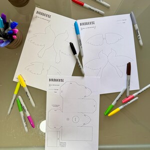 May include: Printable birdhouse craft template with instructions to cut, fold, and glue. The template includes a birdhouse design and two bird shapes. The image also shows various colored markers.
