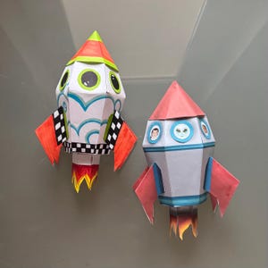 Könnte beinhalten: Zwei bunte Papiermodelle von Raketenschiffen. Eines ist weiß mit blauen Wirbeln, schwarz-weißen Schachflaggen und orangefarbenen und roten Akzenten. Das andere ist grau mit roten Akzenten und einem rosa Oberteil. Beide haben Flammen.