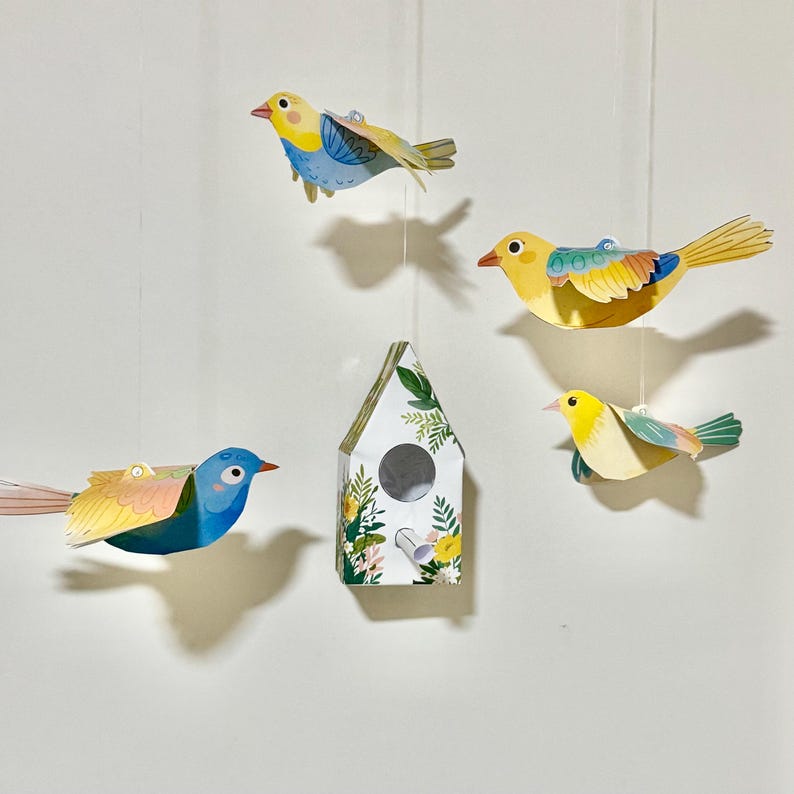 May include: A set of paper bird and birdhouse decorations. The set includes four birds, two yellow with green accents, one blue with yellow accents, and one blue with yellow and pink accents. The birdhouse is white with green and yellow floral accents.