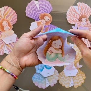 Op de afbeelding: Papieren pop-up kaarten met zeemeerminnen in verschillende kleuren, waaronder roze, paars en koraal. Elke kaart heeft een illustratie van een zeemeermin in een schelp. De kaarten worden in elkaar gezet, wat het 3D-effect laat zien.