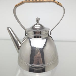 Op de afbeelding: Een theeketel van gepolijst roestvrij staal met een afgerond lichaam, een taps toelopende tuit en een afneembaar deksel met een kleine knop. Het gebogen handvat is omwikkeld met een lichtbruin geweven materiaal voor een textuurrijke grip op de ketel.