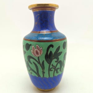 Pode incluir: Um pequeno vaso cloisonné com um corpo verde vibrante decorado com motivos florais pretos e flores de lótus cor-de-rosa. O pescoço é de um azul profundo com detalhes em fio de ouro e a base tem um padrão de ondas azuis e rebordo dourado.