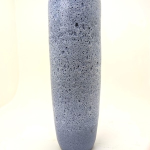 Könnte beinhalten: Eine hohe, zylindrische Keramikvase mit einer texturierten, gesprenkelten Oberfläche in hellblauen und dunkelblauen Farbtönen. Die Oberfläche weist ein poröses, kraterartiges Aussehen mit einem gleichmäßigen, gesprenkelten Muster auf dem gesamten Gefäß auf.