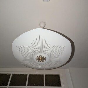 Può includere: Una lampada da soffitto bianca con un ampio paralume in vetro poco profondo a forma di piattino. Il paralume è decorato con un motivo a stella radiante di sottili linee grigie che emanano da un elemento decorativo centrale in metallo dorato.