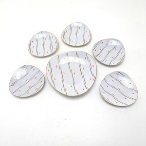 Op de afbeelding: Een set van zes witte keramische schalen met onregelmatige, afgeronde vormen. Elke schaal heeft een herhalend patroon van diagonale gouden lijnen afgewisseld met kleine, verticale rode streepjes. De set bevat één grotere centrale schaal en vijf kleinere schalen.