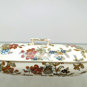 Pode incluir: Uma caixa oval de cerâmica com tampa sobre uma base de cor creme, decorada com um padrão floral multicolorido em tons de vermelho, azul e castanho. A tampa inclui uma pequena pega retangular centrada no topo, a condizer com o design floral.