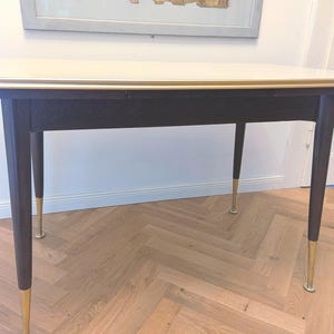 Könnte beinhalten: Ein rechteckiger Esstisch mit einer glatten, cremeweißen Tischplatte und einer dünnen goldfarbenen Metallkante. Der Tisch steht auf vier dunkelbraunen Holzbeinen, die sich nach unten verjüngen und jeweils einen goldfarbenen Metallfuß haben.
