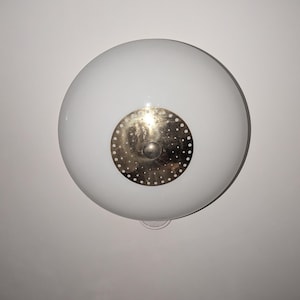 Può includere: Una plafoniera circolare in vetro bianco con un disco centrale in metallo dorato. Il disco dorato è traforato con una serie di piccoli fori equidistanti, disposti in un motivo circolare attorno a un punto centrale liscio e pieno.