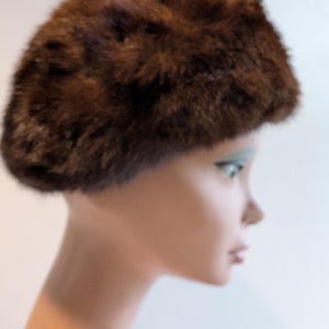 May include: A brown fur hat with a rounded crown. The hat is displayed on a mannequin head. The fur appears to be a mix of dark and light brown shades, creating a textured appearance. The hat is a vintage style.