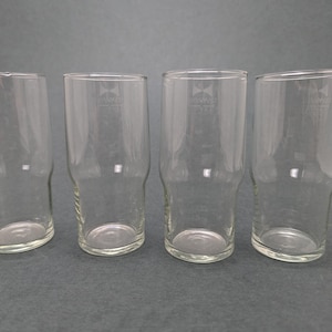 Könnte beinhalten: Vier klare, zylindrische Biergläser mit leicht ausgestelltem Boden. Jedes Glas hat ein Logo oben und eine Messmarkierung. Die Gläser sind in einer Reihe auf neutralem Hintergrund angeordnet.