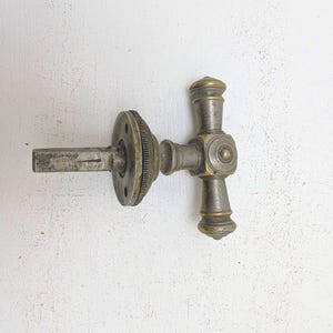 May include: A metallic T-shaped door handle featuring a weathered brass finish. The handle includes a central horizontal bar with rounded decorative ends, a circular mounting plate with a ridged edge, and a square-shaped spindle extending outward.