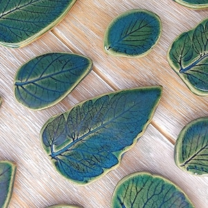 May include: A set of ten ceramic leaf-shaped coasters in shades of green and blue. The coasters are hand-painted with a detailed leaf design.