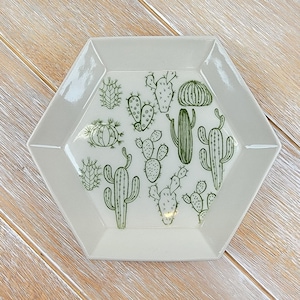 Puede incluir: Un plato hexagonal de cerámica con fondo blanco y un patrón de cactus verde. El plato es perfecto para servir aperitivos o guardar objetos pequeños.
