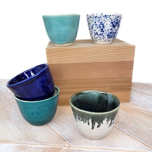 Może przedstawiać: Cztery ceramiczne kubki w różnych kolorach i wzorach. Kubki są ułożone na drewnianym pudełku. Kubki są turkusowe, niebieskie z białymi plamkami, ciemnoniebieskie i zielone z białym szkliwem.