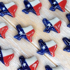 May include: A collection of ceramic Texas-shaped ornaments. Each ornament is painted with the Texas flag design: a blue field with a white star, a white stripe, and a red stripe. The ornaments are arranged on a light wood surface.