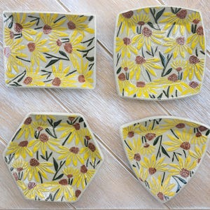 Puede incluir: Cuatro platos decorativos de cerámica de varias formas: cuadrado, rectangular, hexagonal y triangular. Cada plato presenta un diseño de girasol amarillo con centros marrones y hojas verdes sobre un fondo crema.