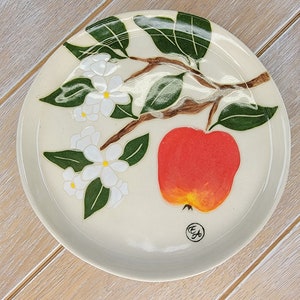 以下が含まれることがあります： 白い陶器の皿に、赤いリンゴと黄色のハイライト、緑の葉、白い花の手描き模様が施されています。皿には「E&F」のサインがあります。