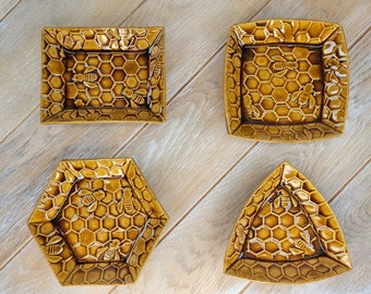 Puede incluir: Cuatro platos de cerámica con un patrón de panal y diseños de abejas. Los platos son de diferentes formas, incluyendo cuadrada, hexagonal y triangular.