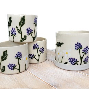 May include: Four ceramic flower pots with white backgrounds and floral designs. The pots feature purple and green flowers with white accents. Three of the pots are stacked on top of each other, while the fourth pot is on a white surface.