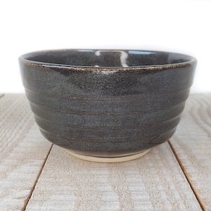 May include: A dark gray ceramic bowl with horizontal textured lines. The bowl has a slightly flared rim and a light beige base. The bowl is sitting on a light-colored wooden surface.