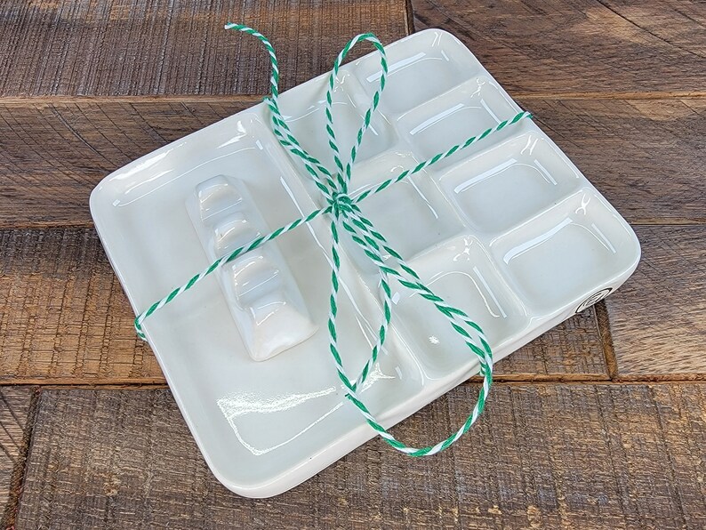 May include: A white ceramic paint palette with nine separate wells, including a long, rectangular well. The palette is tied with green and white twine.