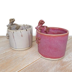 Può includere: Due tazze in ceramica con manici a forma di rana. La tazza a sinistra è bianca con macchie nere e una glassa grigio chiaro. La tazza a destra è rosa intenso con una glassa screziata.