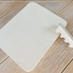May include: A white ceramic rectangular serving tray with a white ceramic handle with four finger holes. The tray is on a light wood surface.
