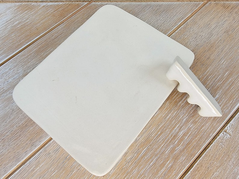 May include: A white ceramic rectangular serving tray with a white ceramic handle with four finger holes. The tray is on a light wood surface.
