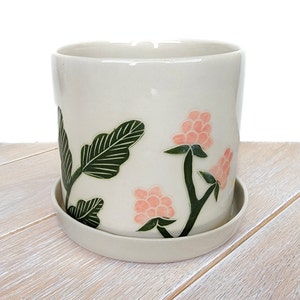 Puede incluir: Maceta de cerámica blanca con un diseño de hojas verdes y acentos de flores rosas. La maceta tiene un platillo a juego.