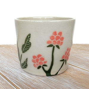 Puede incluir: Una taza de cerámica blanca con un diseño floral. La taza presenta flores rosas con hojas y tallos verdes.
