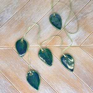 May include: Five glossy, dark green ceramic leaf-shaped ornaments, each featuring a central vein detail and a small hole at the top. Each leaf is individually suspended by a thin, light green ribbon, creating a connected arrangement of hanging decor.
