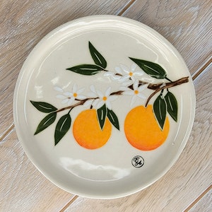 May include: A white ceramic plate with a hand-painted design of a branch of orange blossoms and two oranges. The plate has a brown rim and a small brown mark on the bottom.