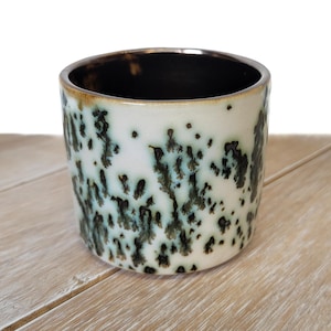 May include: A ceramic cup with a speckled design. The cup has a white base with dark green and black spots. The rim is brown, and the interior is black. The cup sits on a light-colored wooden surface.