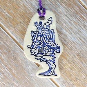 Puede incluir: Un adorno de cerámica blanca con forma de casa con una pierna y un pie esqueléticos. La casa está decorada con detalles azules y blancos, incluyendo un diseño de calavera y huesos cruzados.