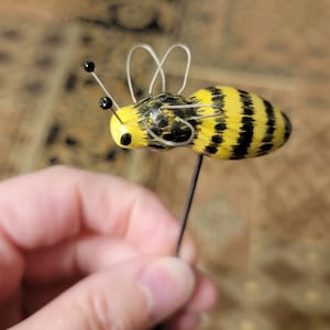 May include: A yellow and black bee figurine with wire antennae and a black bead at the end of each antenna. The bee is attached to a thin black wire.
