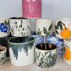 May include: A selection of ceramic mugs with diverse designs and colours. Some mugs display hand-painted illustrations of blueberries, flowers, and oranges. Others have speckled or dripped glaze patterns. The mugs are cylindrical and appear handmade.