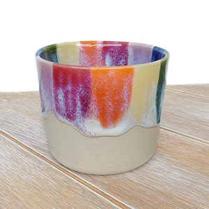 May include: A ceramic mug with a rainbow stripe design. The mug is glazed in a light beige colour with a white drip effect. The stripes are in shades of purple, red, orange, yellow, and blue.