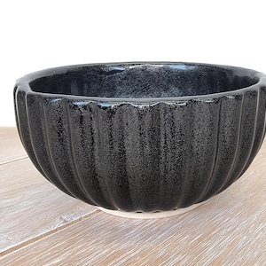 May include: A black ceramic bowl with a textured, ribbed exterior. The bowl has a glossy finish and a white base. The bowl is sitting on a light-coloured wooden surface. The bowl is designed for serving food.