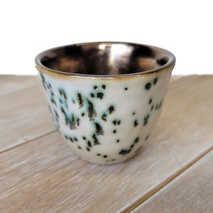 May include: A small ceramic cup with a tapered shape, featuring a creamy white exterior speckled with dark green and black spots. The interior of the cup has a smooth, metallic bronze finish that extends to the rim, creating a contrast.