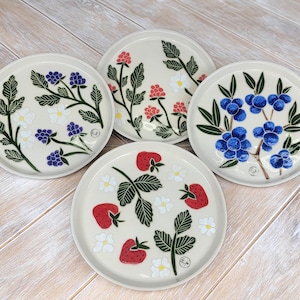 May include: Four ceramic plates with different fruit designs. The plates are white with a cream glaze and feature hand-painted illustrations of strawberries, blueberries, and raspberries. Each plate has a different design and color scheme.