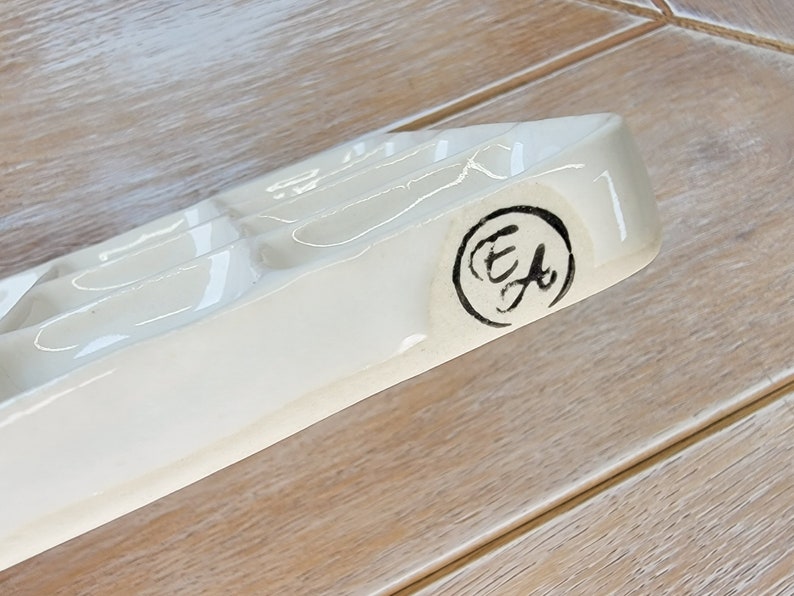 May include: White ceramic paint palette with six wells and a black circular stamp with the letters "E.F." inside.