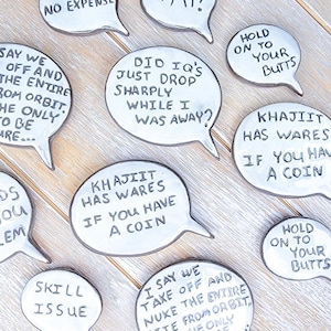 May include: A collection of white, speech bubble-shaped ceramic pieces with black handwritten text. Phrases include "Did IQ's just drop sharply while I was away?" and "Khajiit has wares if you have a coin."