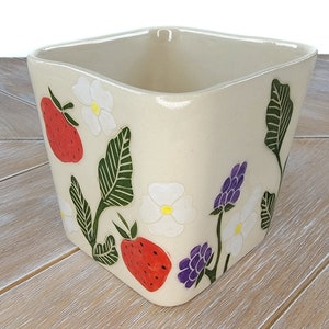 May include: A white ceramic square planter with a floral design. The planter features red strawberries, white flowers, green leaves, and purple flowers.