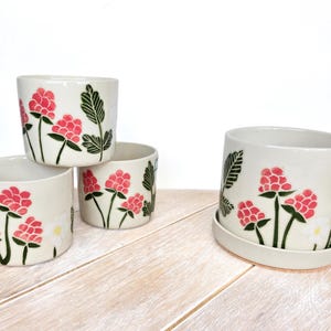 May include: Four off-white ceramic planters featuring hand-painted pink raspberry clusters, dark green leaves, and small white flowers. Three planters are cylindrical, while one larger planter sits on a matching circular ceramic saucer.