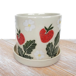 May include: A white ceramic planter with a saucer, featuring a hand-painted design of red strawberries, green leaves, and white flowers.