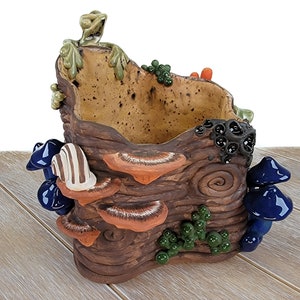 Puede incluir: Una maceta de cerámica con forma de tronco de árbol con una rana en la parte superior. La maceta tiene una superficie marrón texturizada con detalles verdes, azules y naranjas. Hay setas y otros detalles en la maceta.