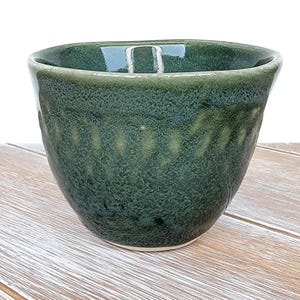 Może przedstawiać: Zielona ceramiczna miska z pstrokatym szkliwem. Miska ma zaokrąglony kształt i lekko rozszerzający się brzeg.